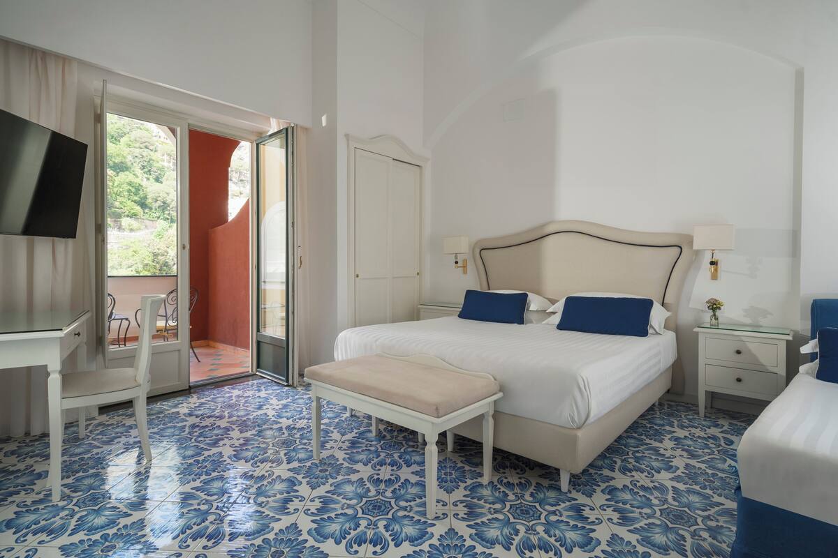 Une chambre d’hôtel comprenant un lit, un bureau, une chaise, un banc, une télévision et une porte donnant sur un balcon.