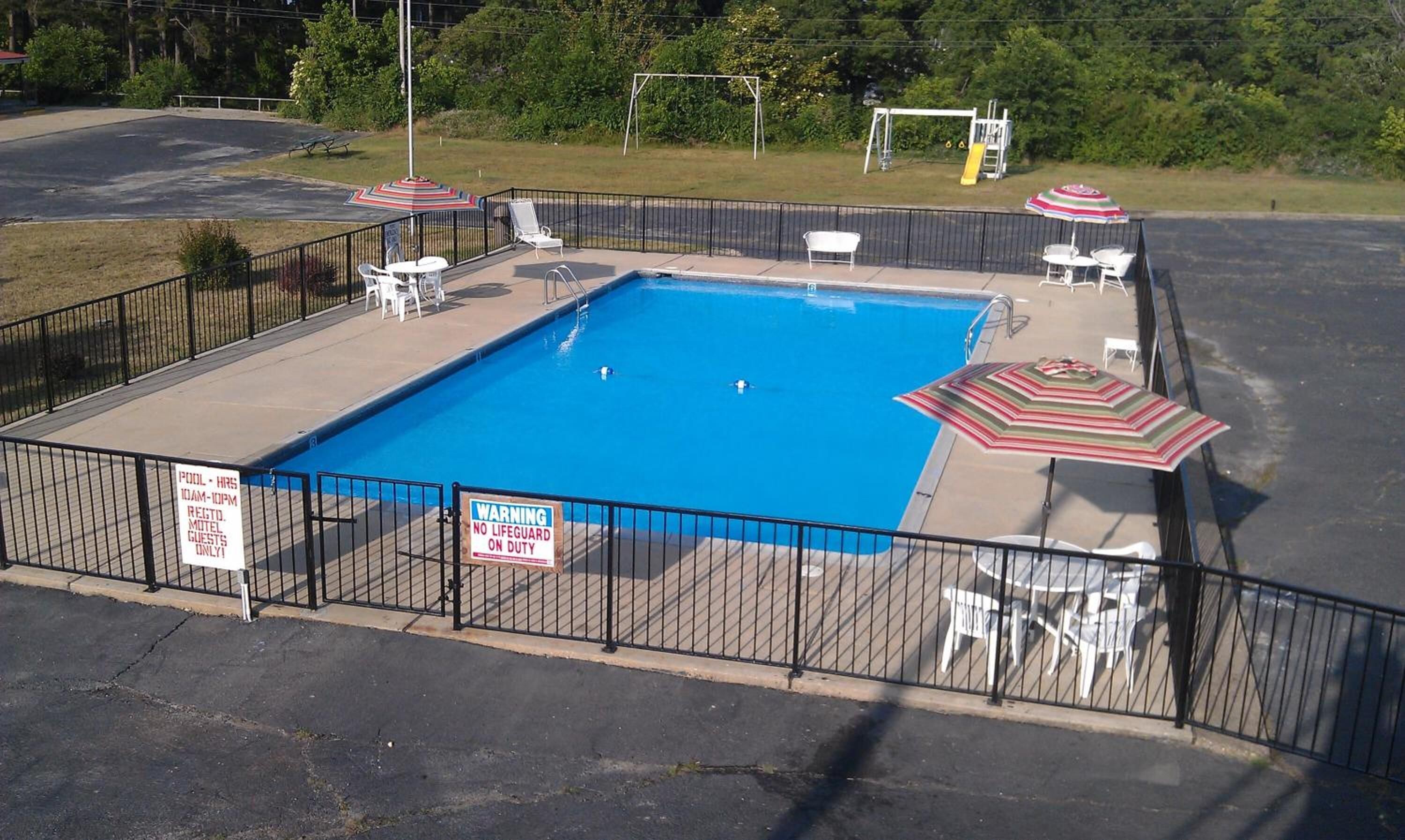 A rectangular pool with a fenced area, surrounded by lounge chairs and umbrellas. Signs indicate the pool hours and a warning about no lifeguard on duty.