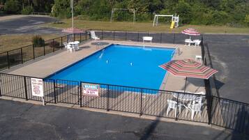 A rectangular pool with a fenced area, surrounded by lounge chairs and umbrellas. Signs indicate the pool hours and a warning about no lifeguard on duty.