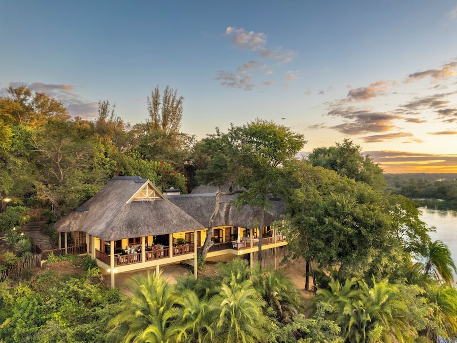 A thatched-roof building with outdoor seating, surrounded by lush greenery and a river.