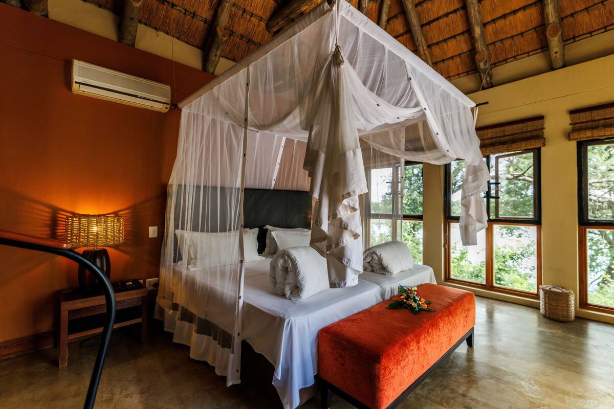 A room with a four-poster bed, a bench, a lamp, and large windows.