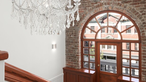 A staircase with a wooden handrail and white balusters, leading to a room with a large arched window and a chandelier. - Victorian Hotel (Vancouver)