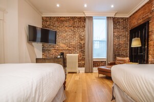 A hotel room with two beds, a brick wall, a TV, a radiator, a window with curtains, and a leather chair.
