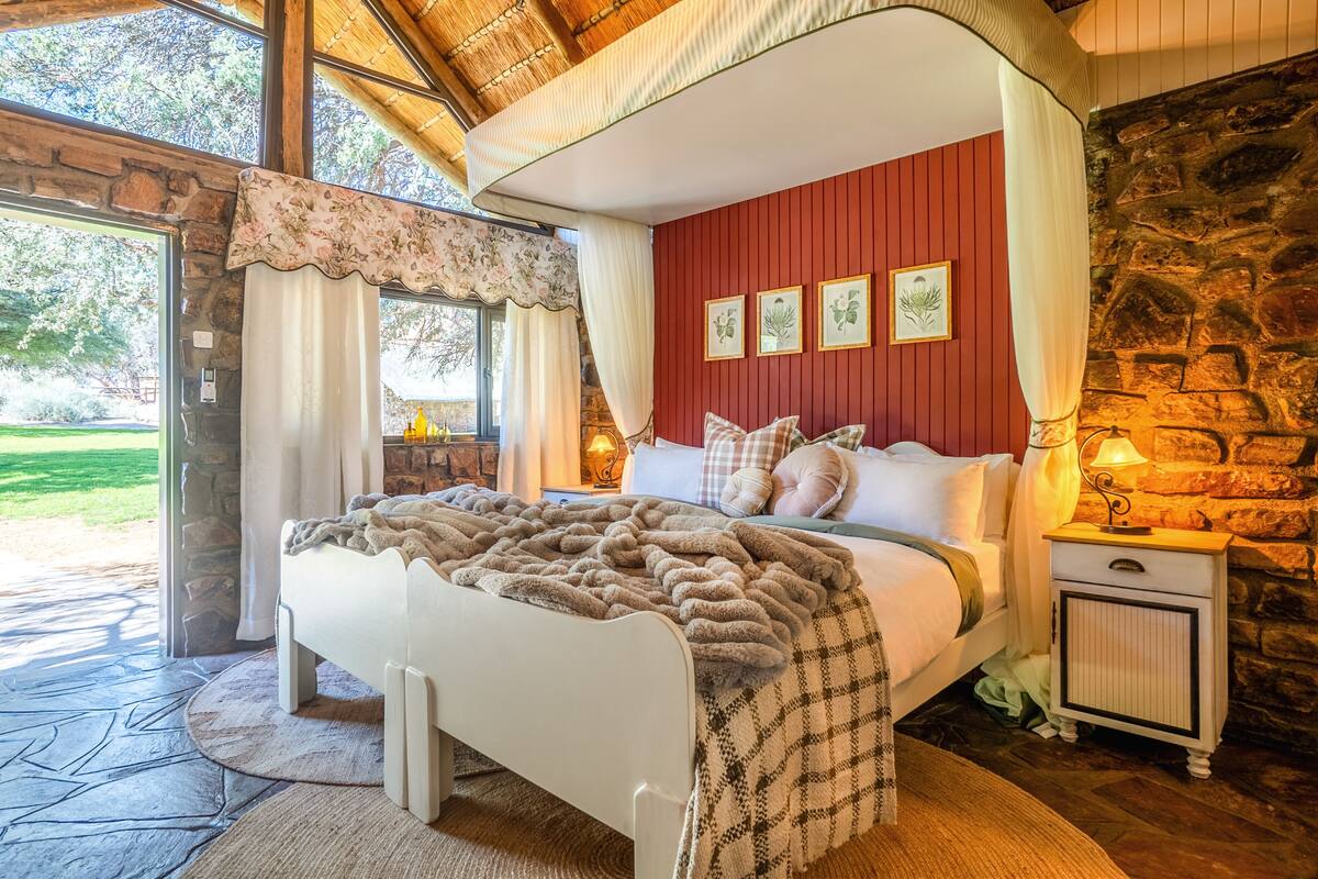 Une chambre avec un mur en pierre, un lit recouvert d’une couverture à carreaux, une table de chevet avec une lampe et une fenêtre avec des rideaux.
