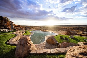 Zona de piscina ajardinada con senderos de piedra, rocas grandes y sillones de descanso.