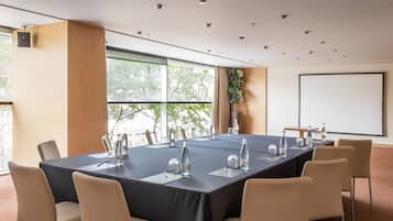 Un salón de conferencias con una larga mesa, sillas, una pantalla de proyector y un amplio ventanal con vistas a árboles.