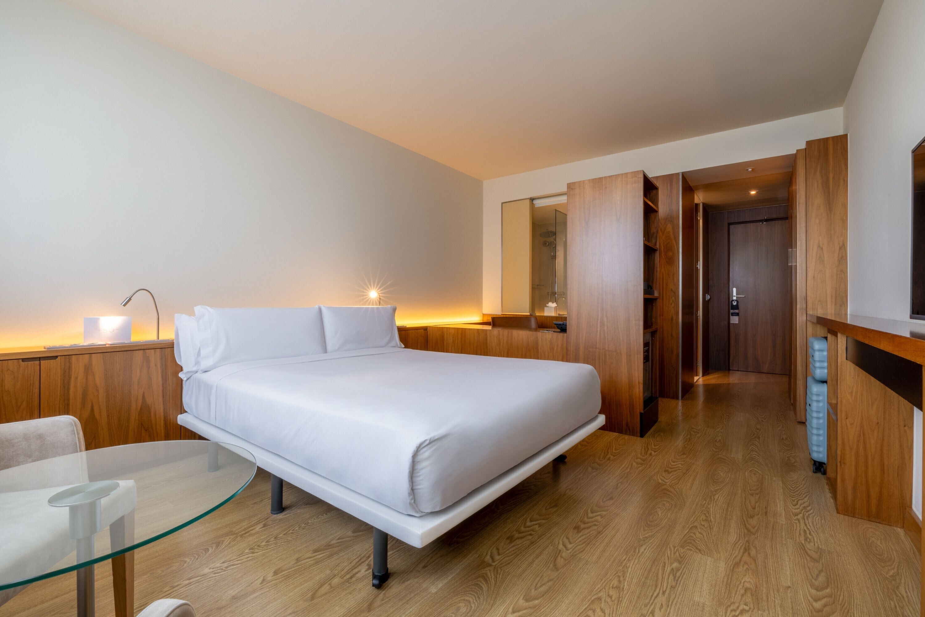 Quarto de hotel moderno com cama, mesa de vidro, cadeira, escrivaninha e espelho.