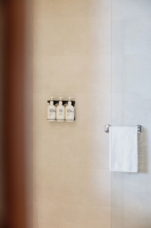 A bathroom with a beige tiled wall, a white towel hanging on a metal towel rack, and three white bottles of shampoo and conditioner on a black shelf. - Hotel SB Diagonal Zero 4 Sup (Barcelona)