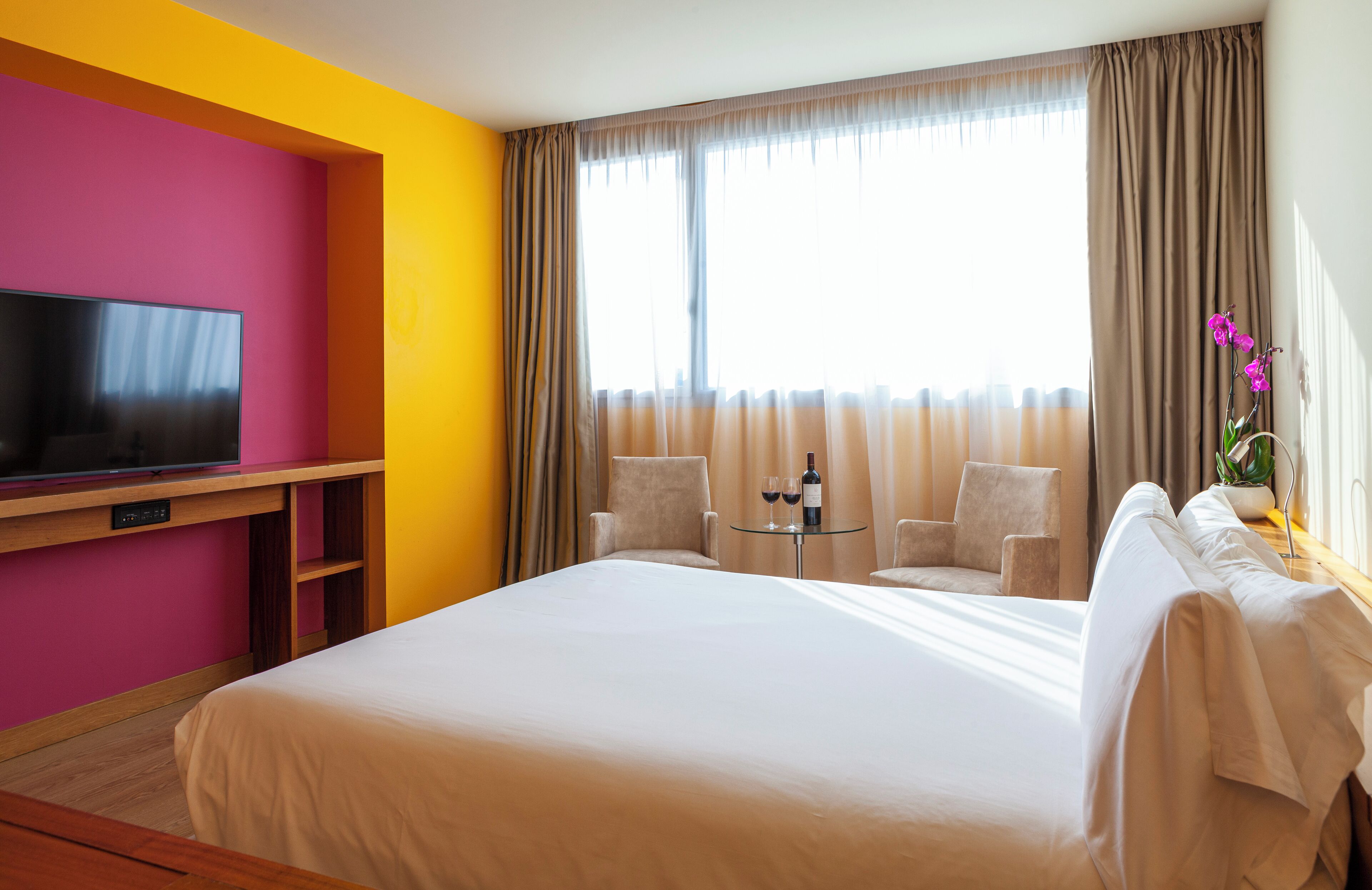 Quarto de hotel com uma cama grande, televisão sobre um suporte de madeira, duas cadeiras, uma mesa pequena com uma garrafa e copos, e uma janela com cortinas.
