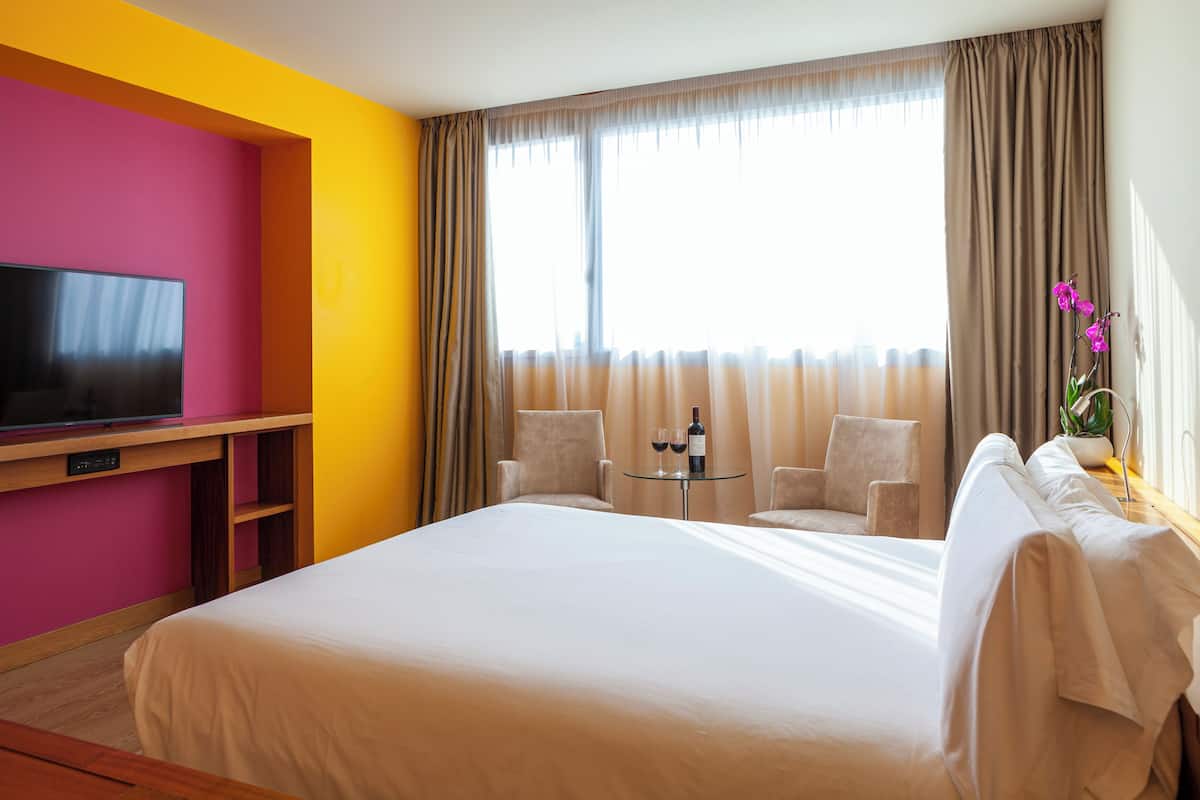 Une chambre d’hôtel avec un grand lit, un téléviseur posé sur un meuble en bois, deux chaises, une petite table avec une bouteille et des verres, et une fenêtre avec des rideaux.