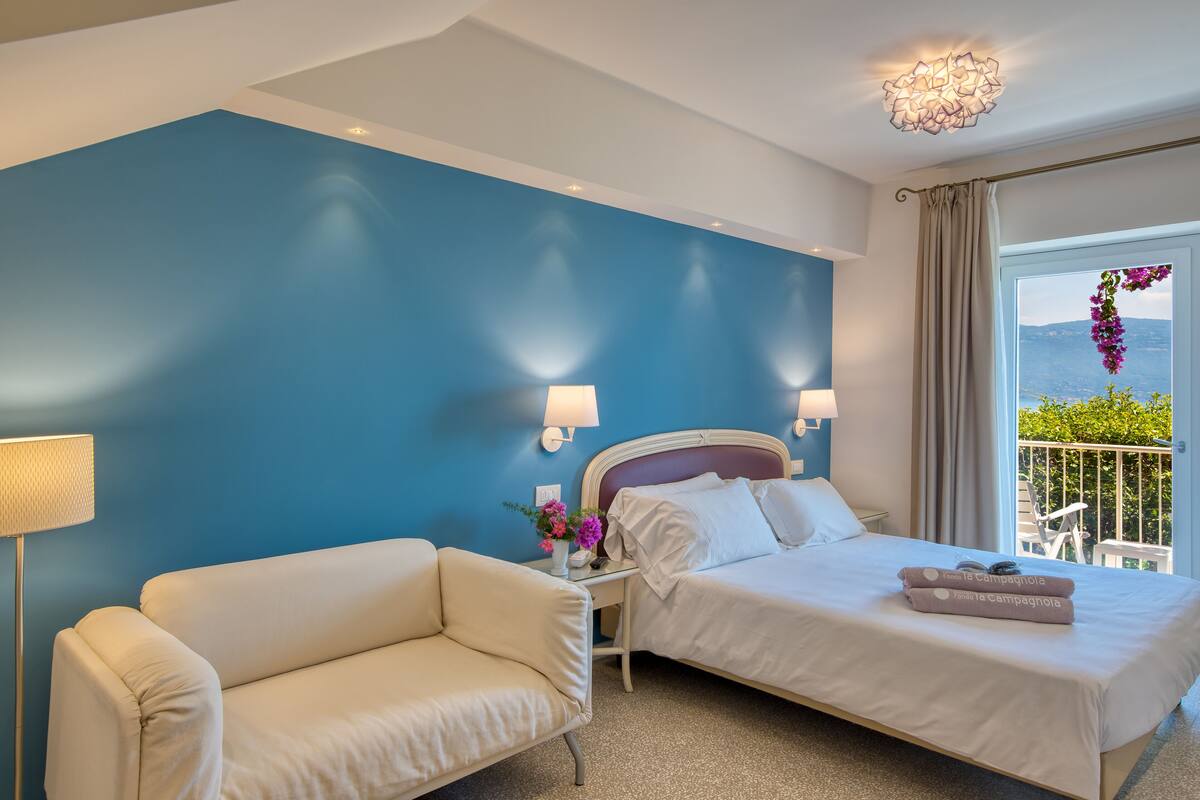 Ein Hotelzimmer mit blauer Akzentwand, einem Bett mit weißen Bettwäsche, einem weißen Sessel, einem Nachttisch mit Lampe und Blick ins Freie.