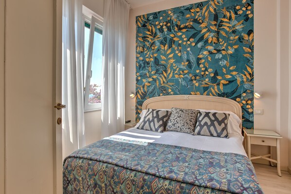 A bedroom with a patterned bedspread, a headboard with a floral tapestry, and a bedside table with a lamp.