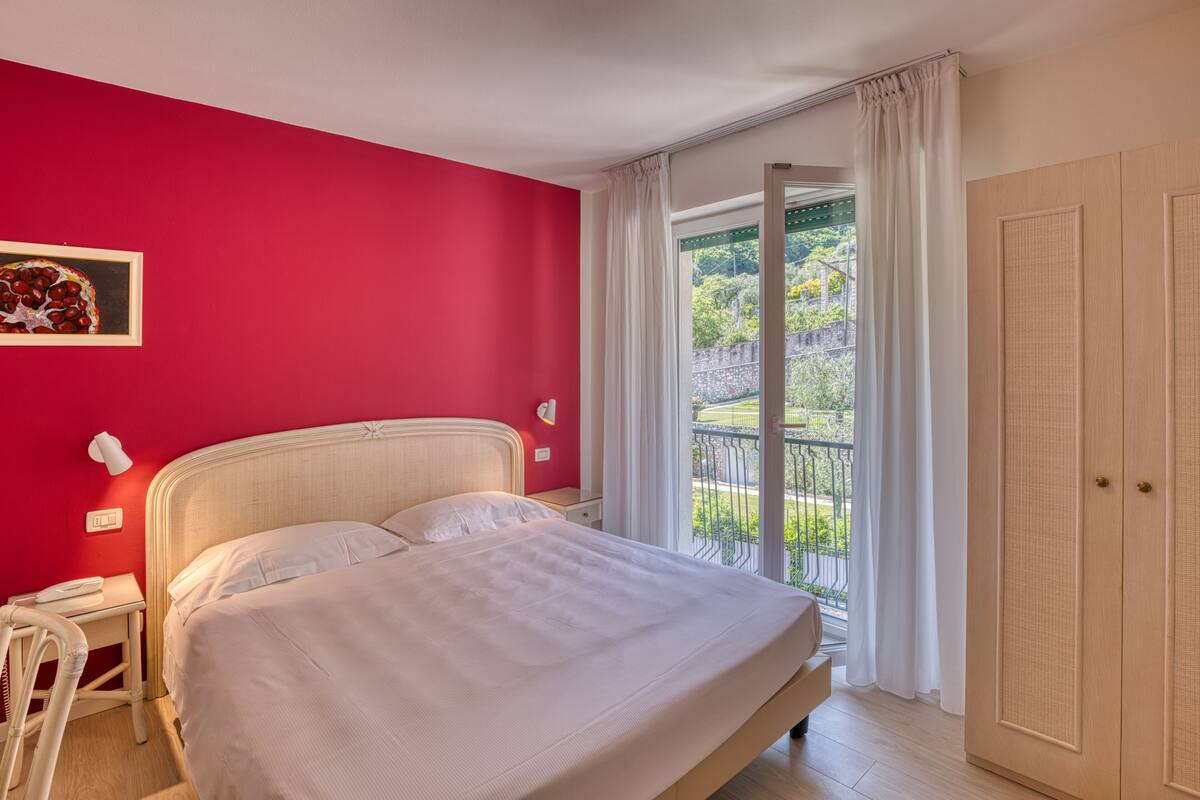 Ein Schlafzimmer mit einem großen Bett, einer roten Wand, einem Fenster mit Blick auf Grünflächen und einer Tür.