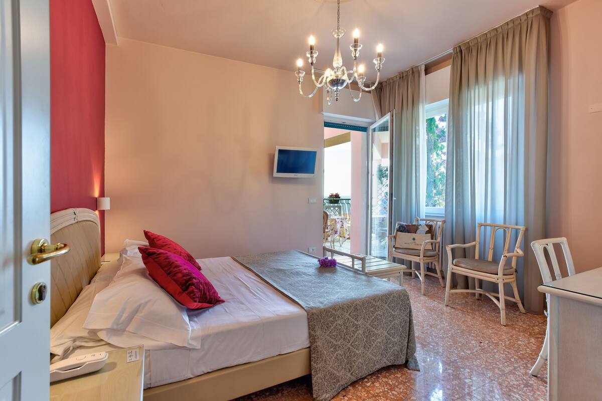 Ein Schlafzimmer mit Bett, Nachttisch, Sessel, Schreibtisch, Fernseher und einem Balkon mit Tisch und Stühlen.