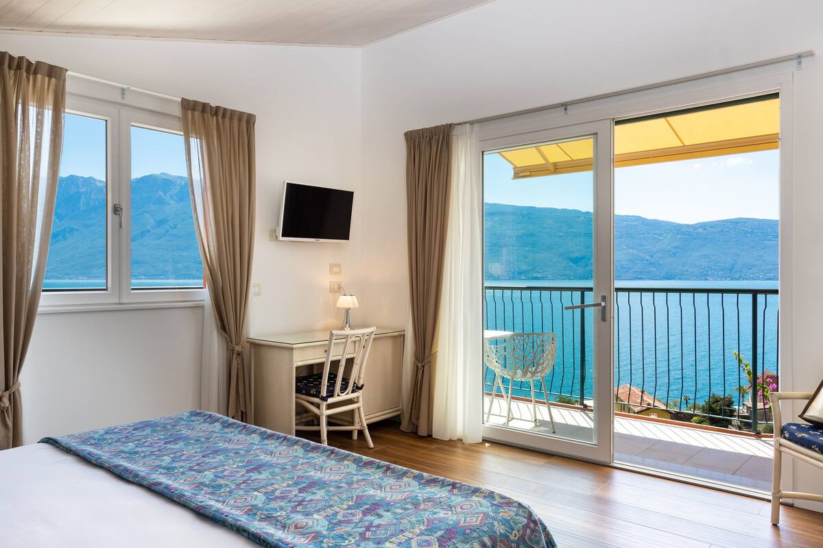 Ein Schlafzimmer mit Bett, Schreibtisch, Stuhl und einem Balkon mit Blick auf das Meer und die Berge.