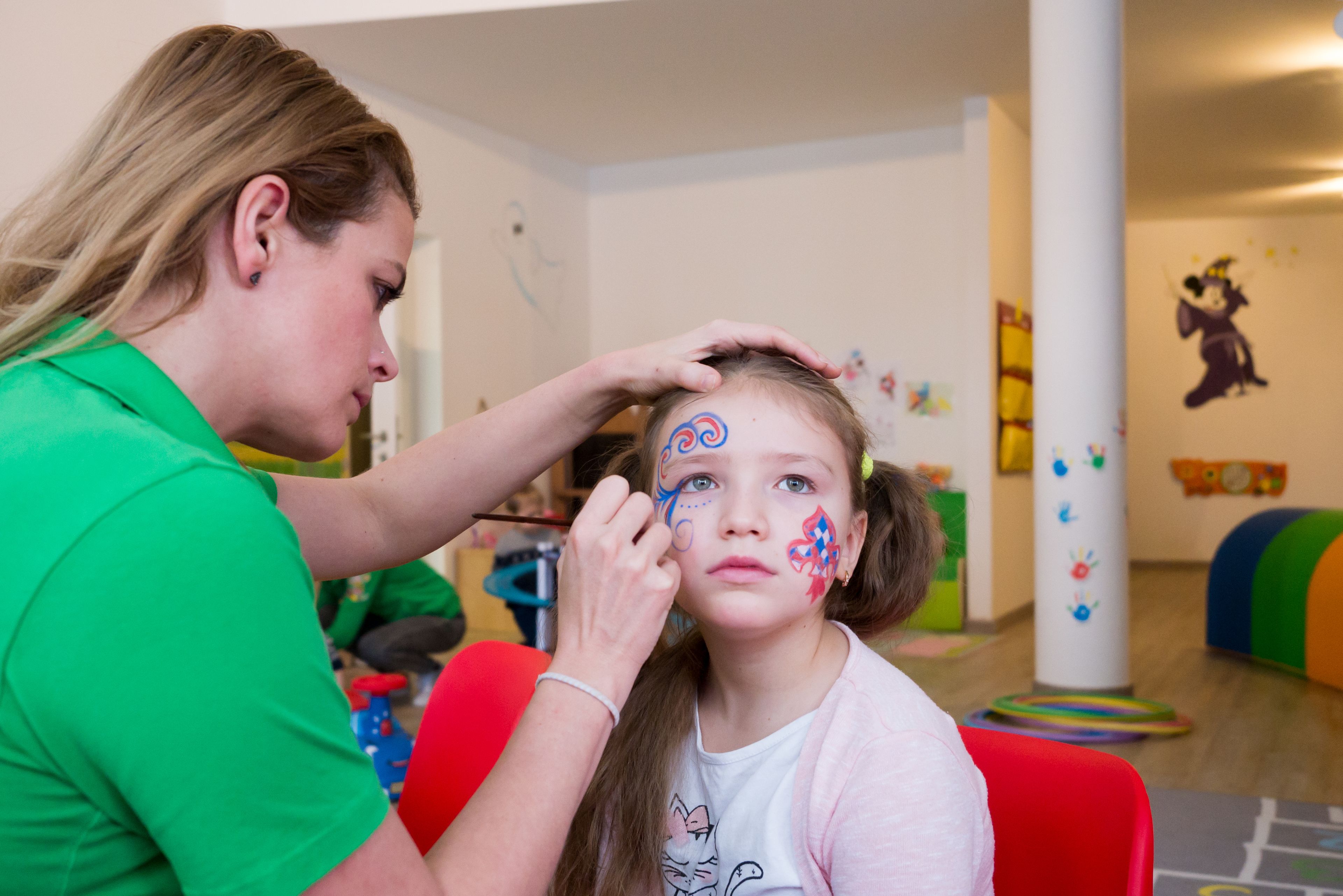 A child with face paint being attended to by an adult in a room with colorful decorations and a play area.