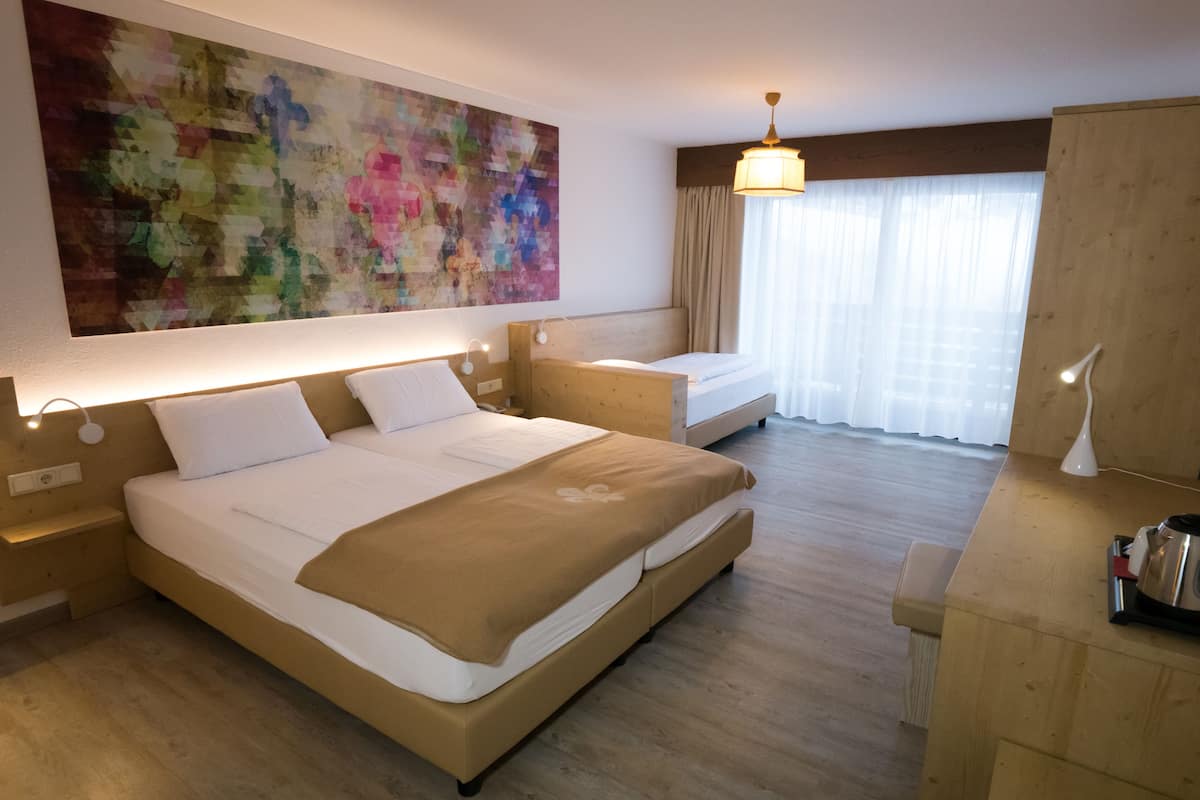 Una habitación de hotel con una cama grande, una cama más pequeña, un escritorio, una silla, una cafetera y un cuadro abstracto de gran tamaño en la pared.
