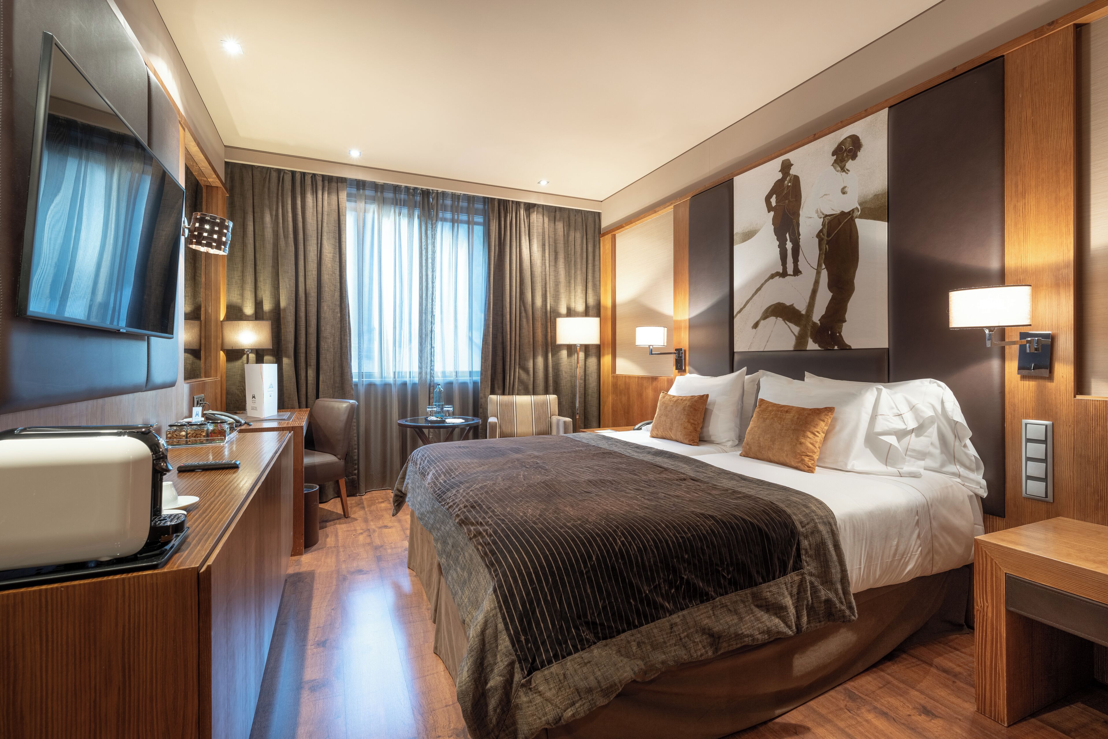 Una habitación de hotel moderna con una cama grande, un escritorio con computadora, una silla, un televisor y un cuadro en la pared.