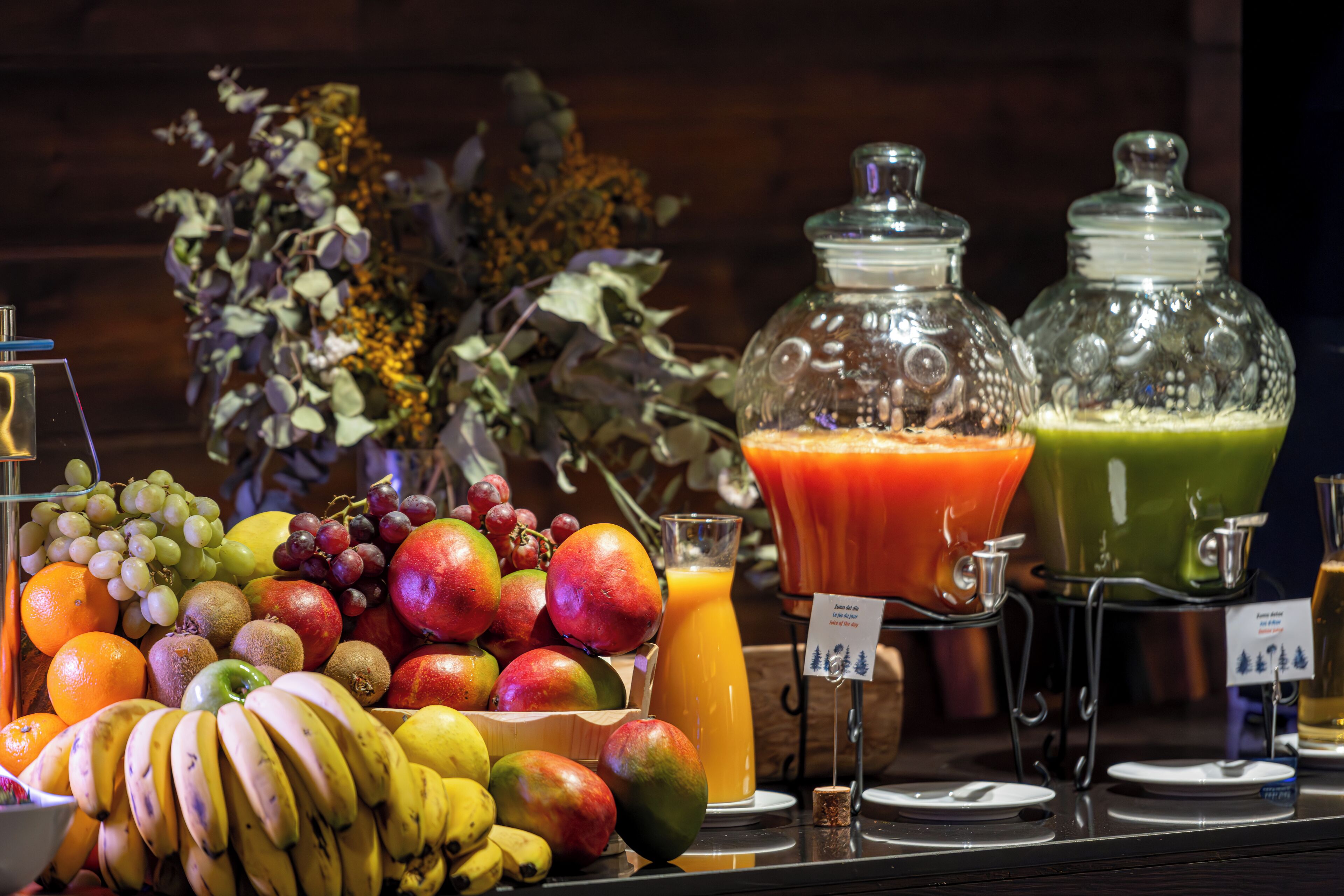 Une présentation de fruits et de jus, avec divers fruits, un verre de jus d’orange et deux bocaux en verre contenant des jus de différentes couleurs.