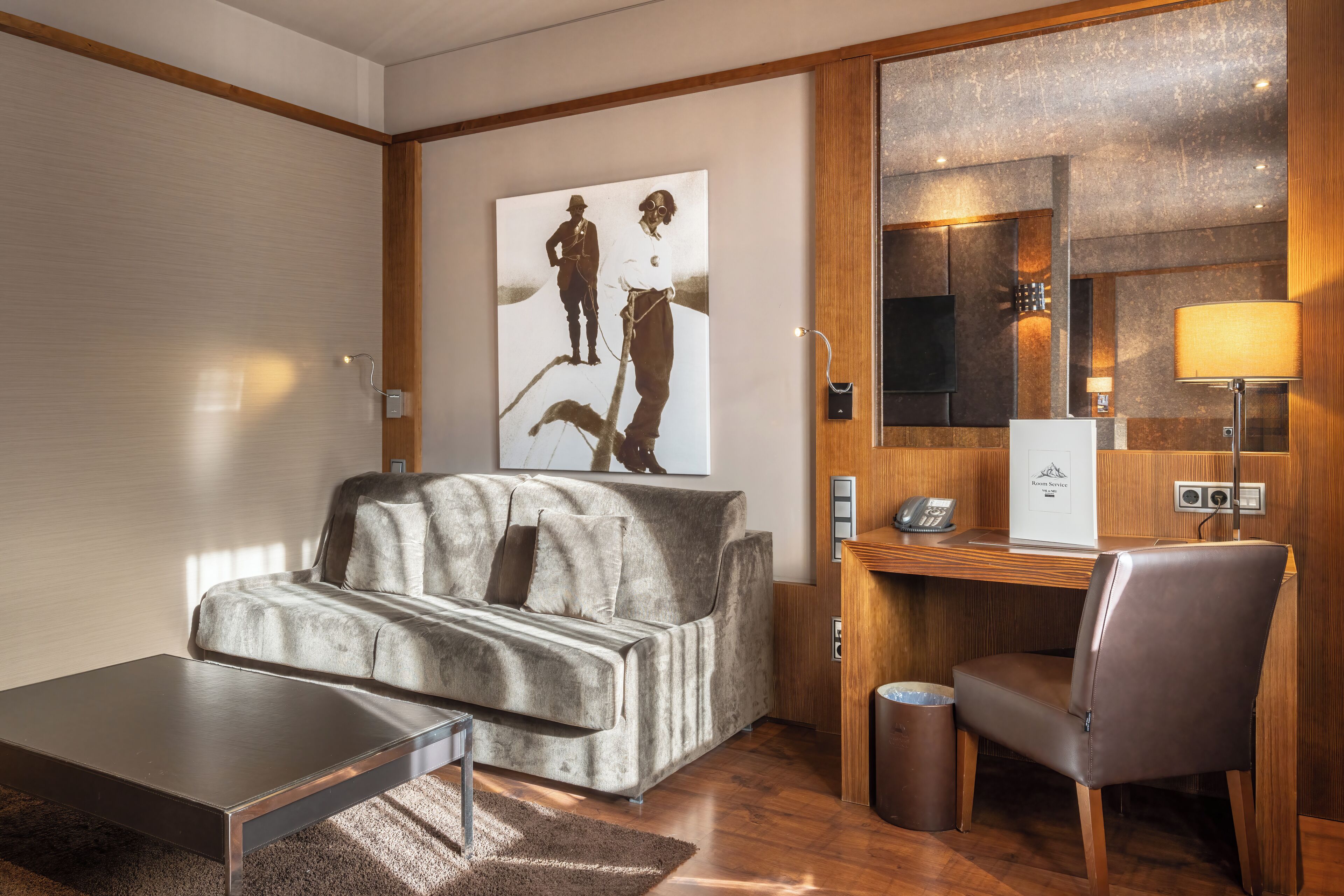 A modern hotel room with a sofa, a small table, a chair, a trash bin, a painting on the wall, a desk with a telephone, and a mirror.