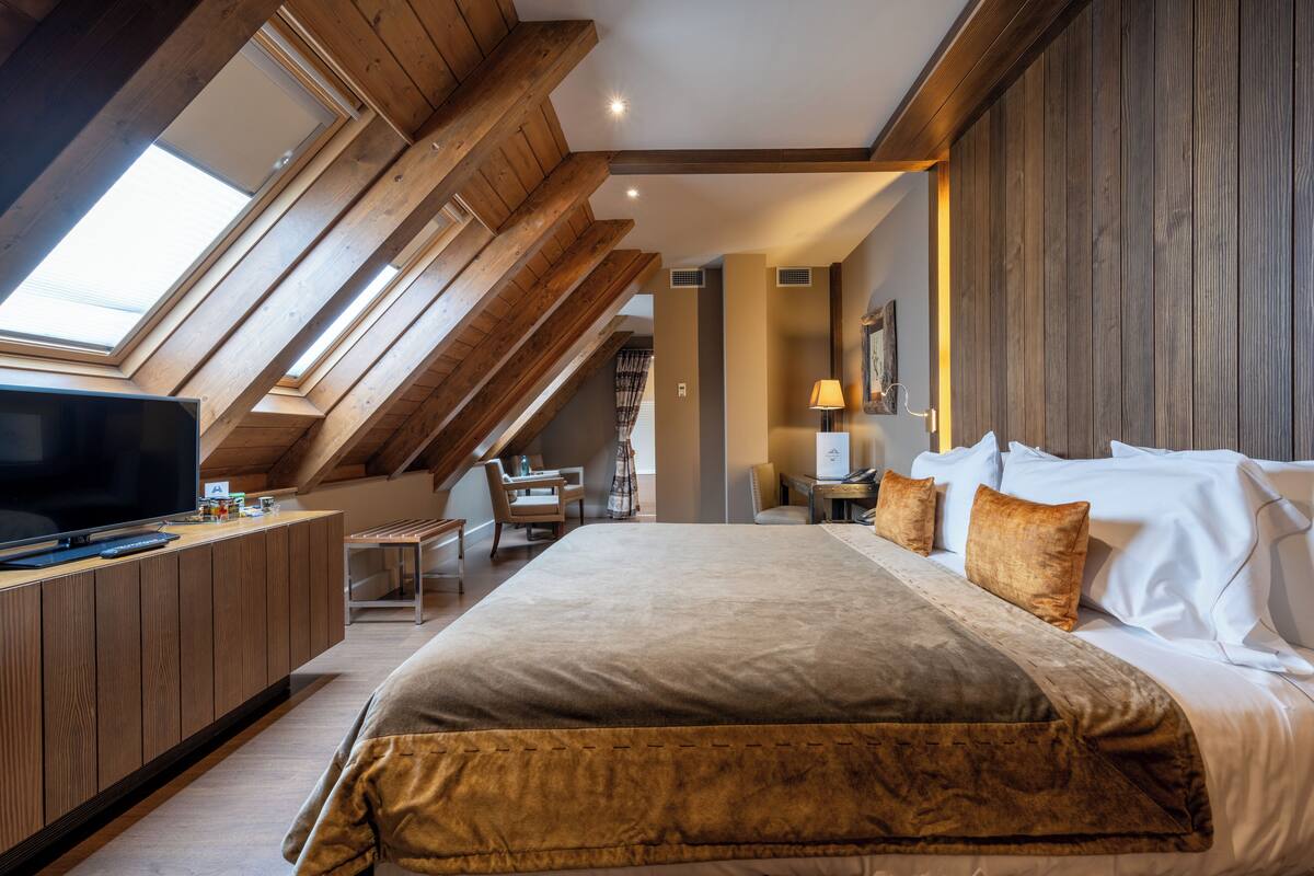 A bedroom with a large bed, wooden ceiling beams, a TV, and a desk with a chair.