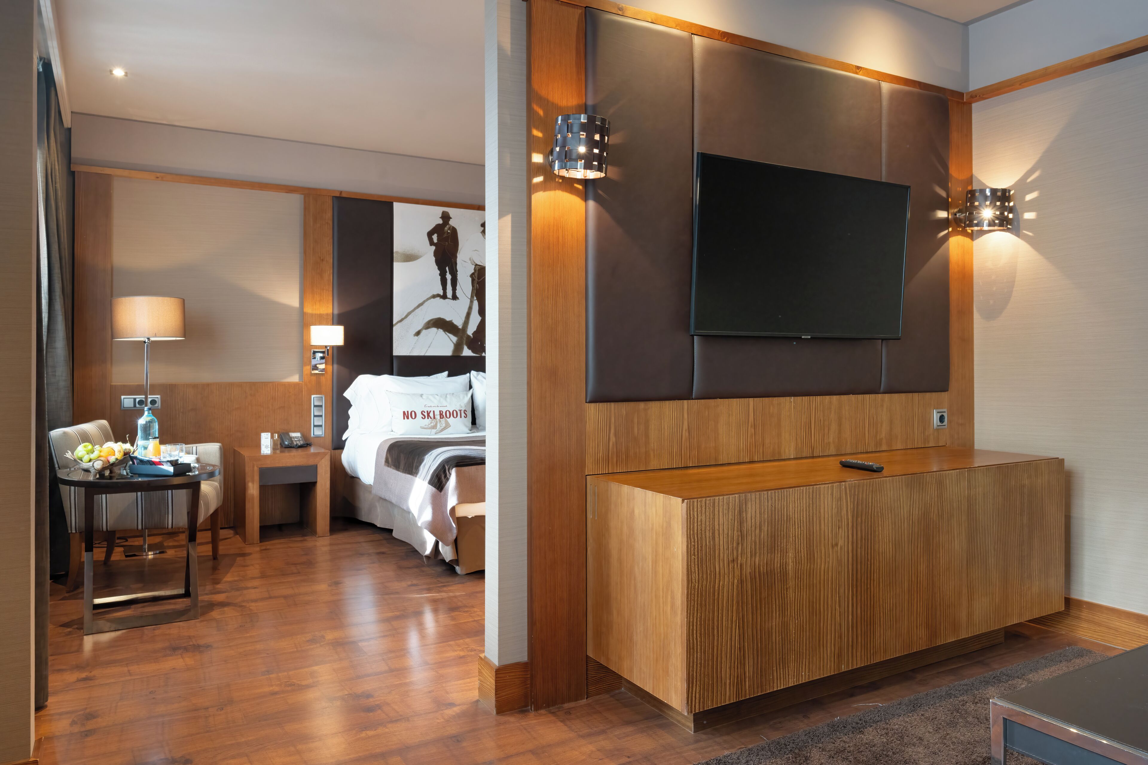 Une chambre d’hôtel avec un plancher en bois, un lit recouvert d’une couette blanche, un bureau avec une lampe, un téléviseur fixé au mur et un luminaire mural.
