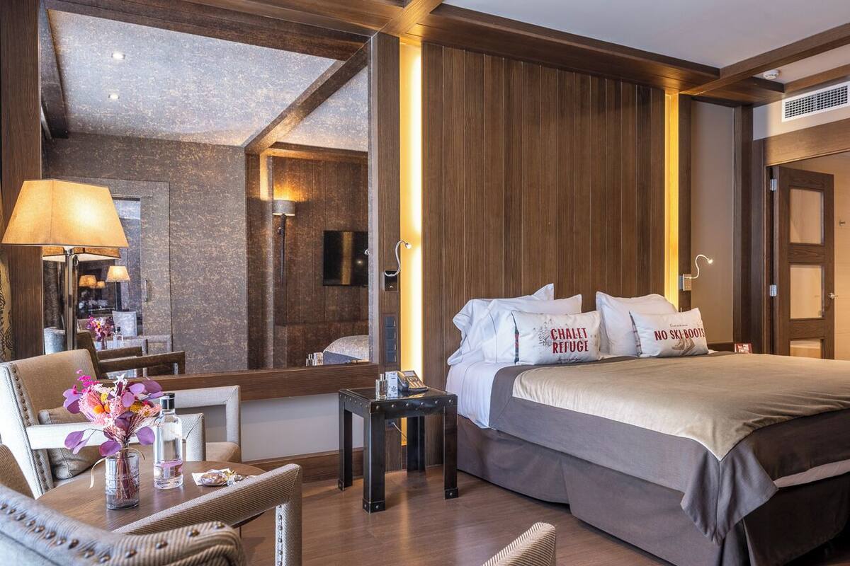 A modern hotel room with a large bed, a sitting area with a table and chairs, a mirror, and a wooden accent wall.
