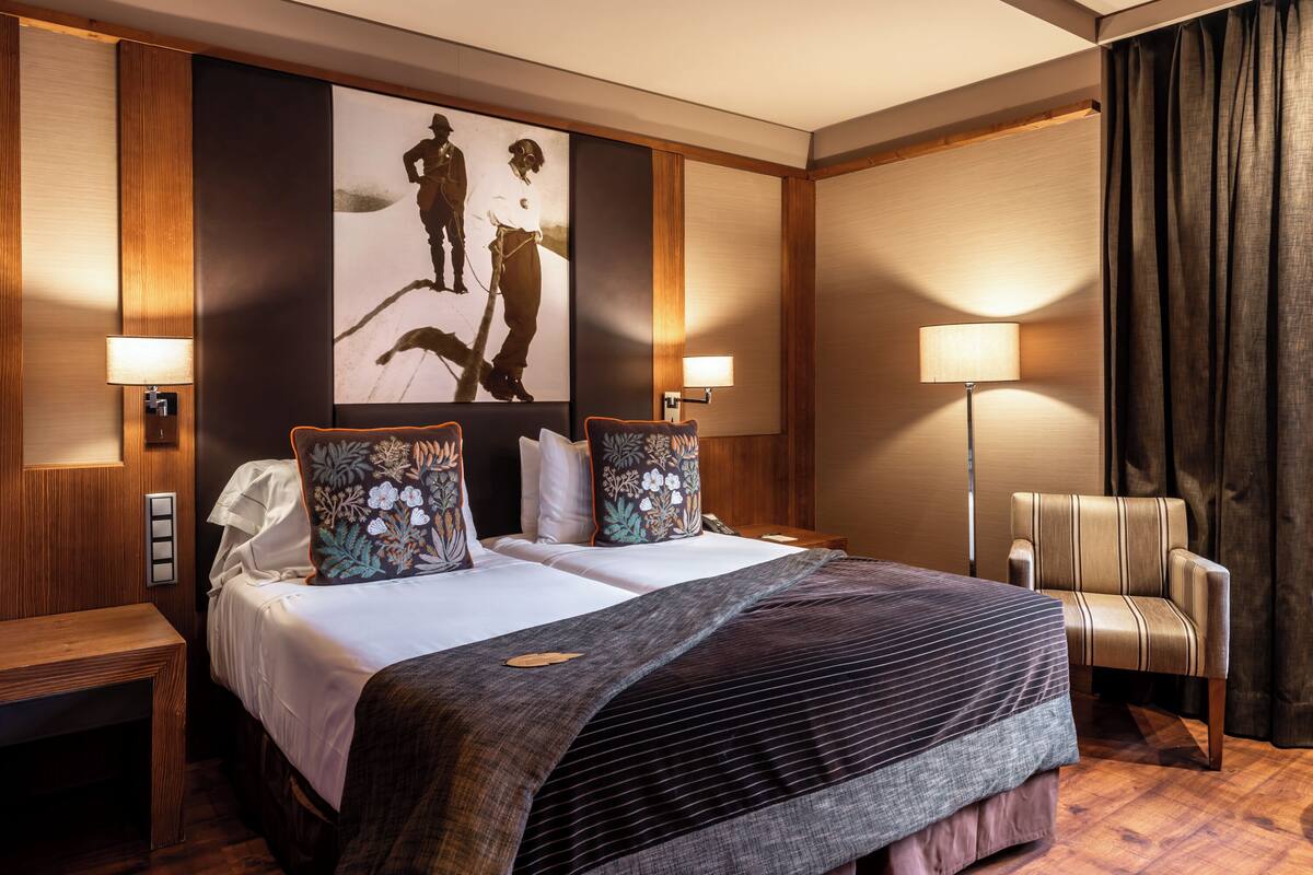 A hotel room with a large bed, two bedside lamps, a wooden bench, a chair, and a large painting on the wall.