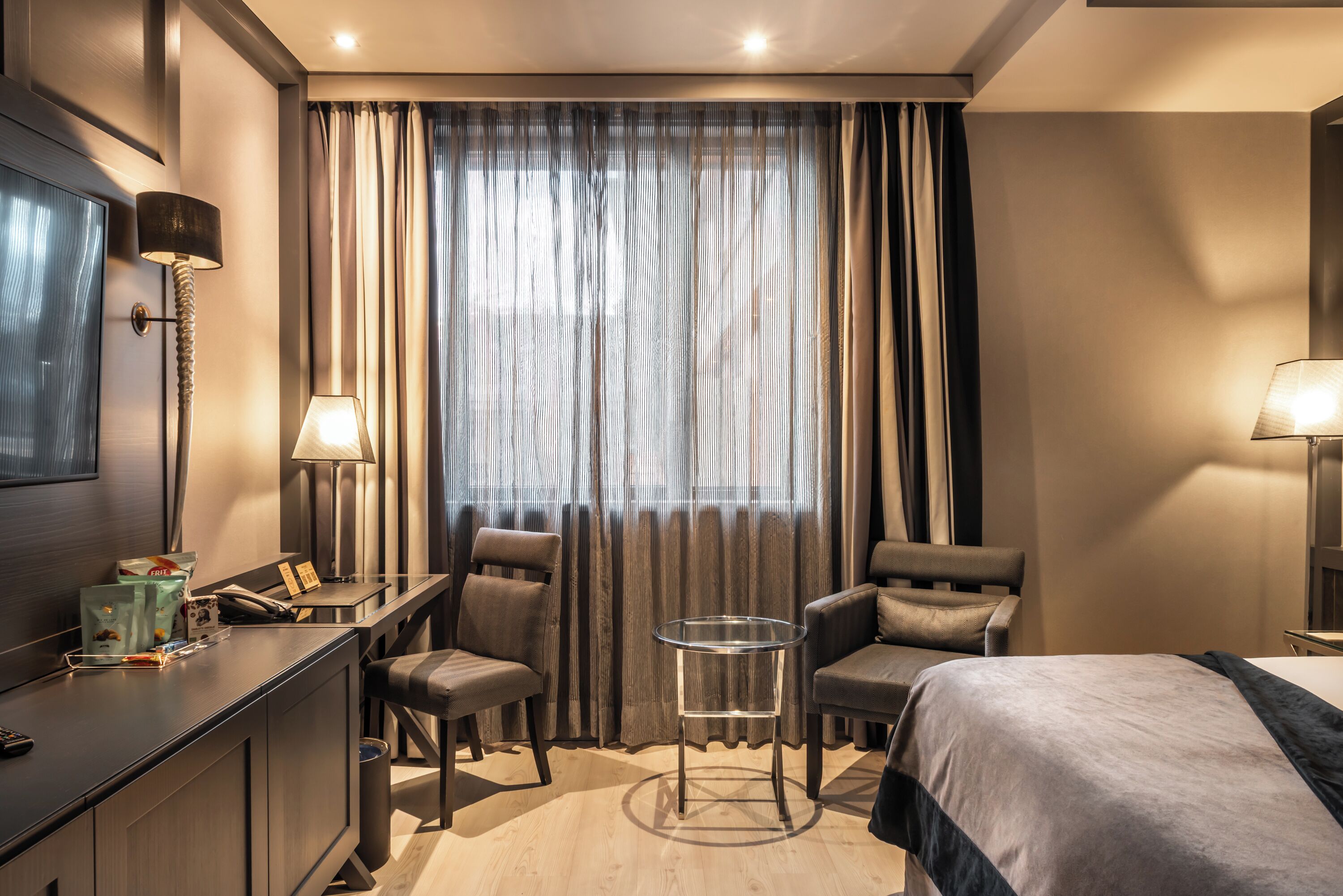 A hotel room with a desk, two chairs, a small table, a bed, and a large window with curtains.