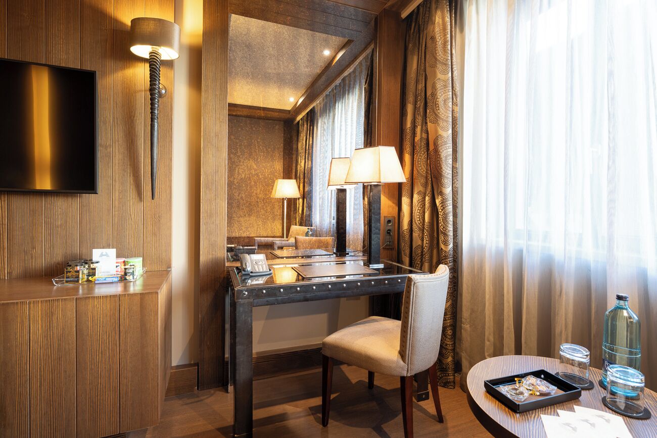 Une chambre d’hôtel avec un bureau en bois, une chaise, un téléviseur à écran plat, deux lampes, une petite table avec un plateau, et une grande fenêtre avec des rideaux.