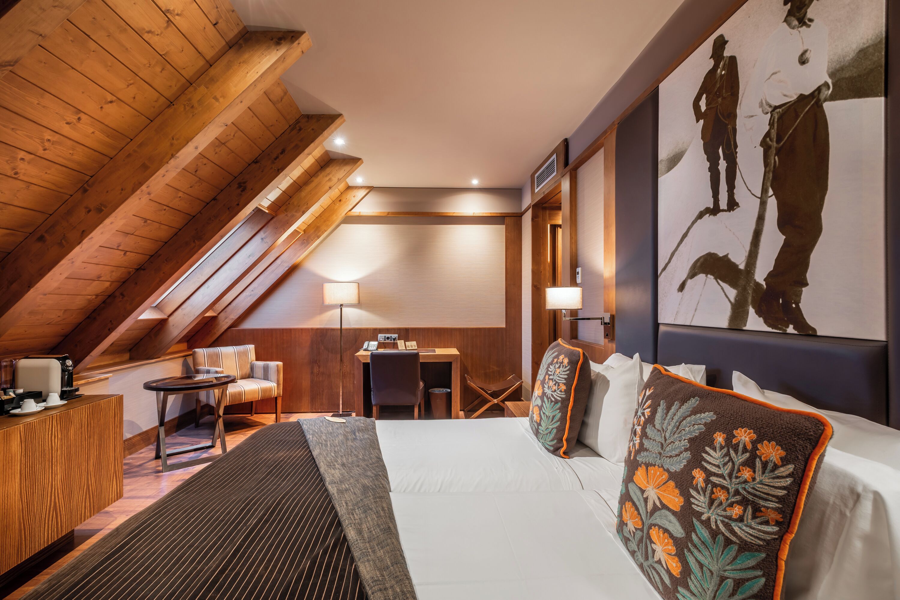 Una camera d'albergo con soffitto a falde, un letto con cuscini floreali, una scrivania con una sedia e un quadro appeso al muro.