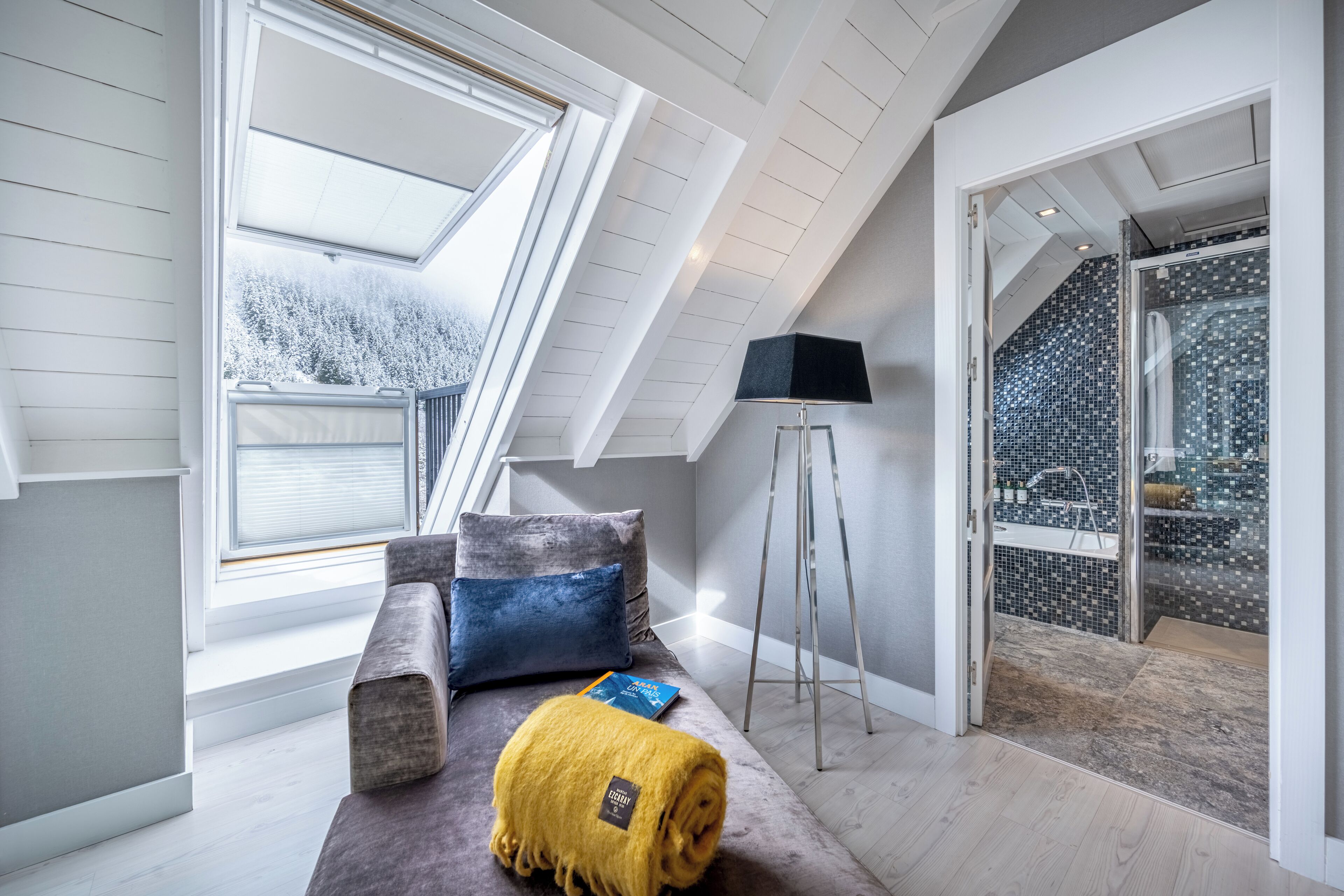 A modern loft bedroom with a sloped ceiling, a cozy seating area with a sofa and cushions, a standing lamp, and a view of a snowy landscape through a large window.