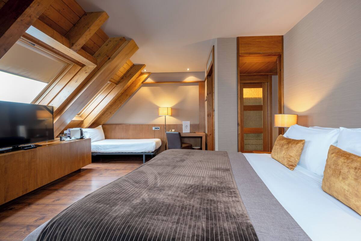 A hotel room with a large bed, a desk, a television, and a window with wooden beams.