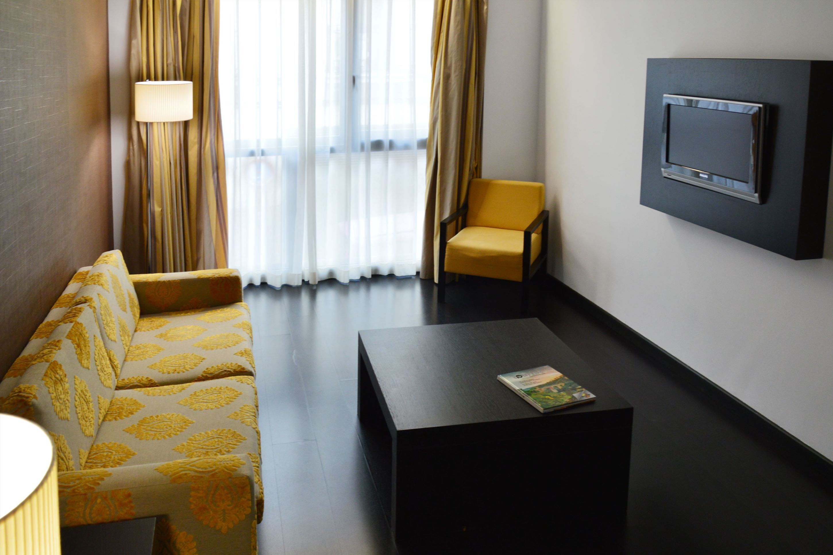 A modern hotel room with a sofa, a chair, a coffee table, and a mounted TV.