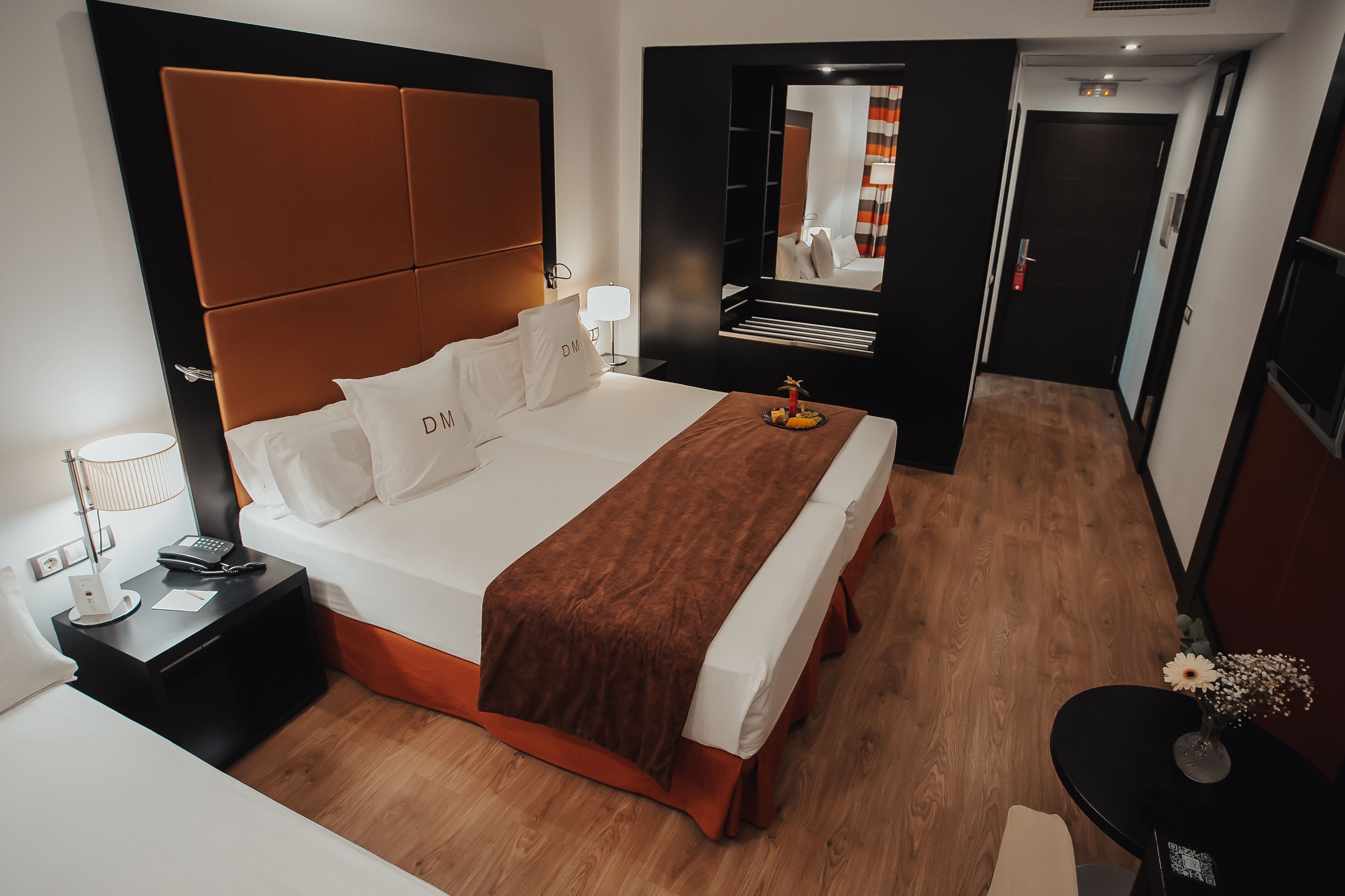 A hotel room with a large bed, a bedside table with a lamp, a small table with a vase, and a mirror on the wall.