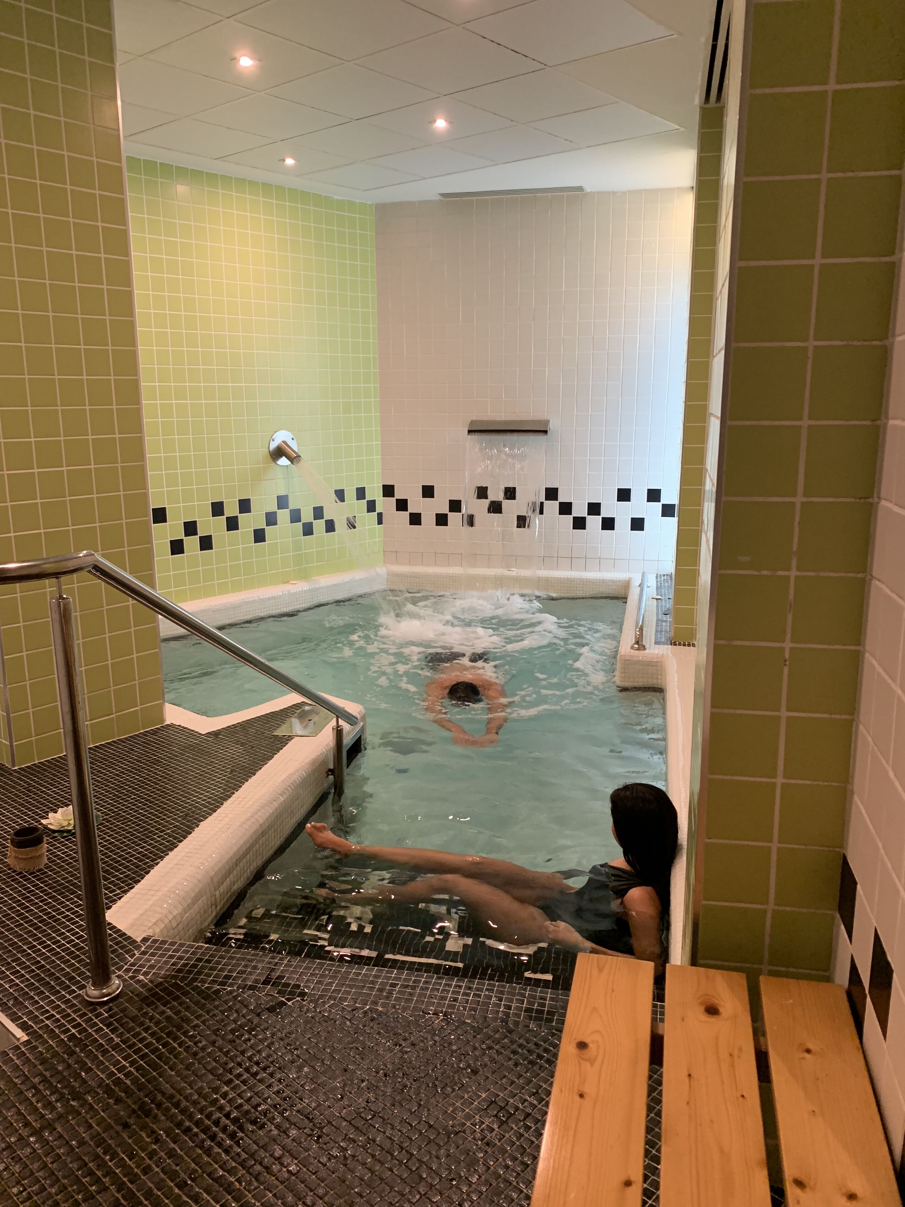 Zona de piscina con un muro de azulejos, una fuente de agua y un banco de madera.
