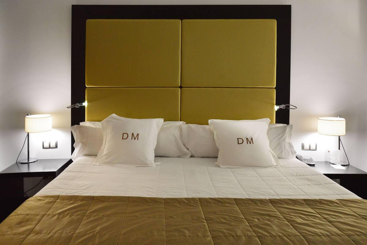 A hotel room with a large bed, two bedside tables with lamps, and a headboard with the initials "DM".