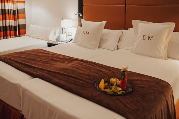 Habitación de hotel con una cama grande, ropa de cama blanca y almohadas con las iniciales 'DM'. Un plato de fruta está sobre la cama.