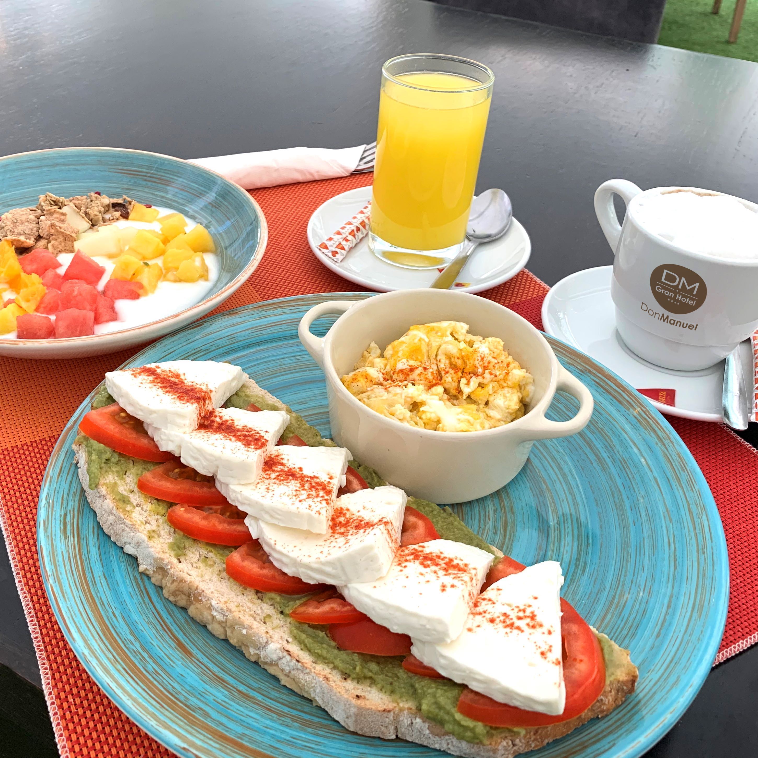A breakfast setting with toast topped with tomatoes, cheese, and guacamole, a bowl of scrambled eggs, and a glass of orange juice.