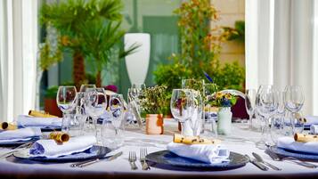 Una mesa dispuesta para una comida, con varias copas de vino, platos y cubiertos, rodeada de plantas en macetas y un jarrón con flores.