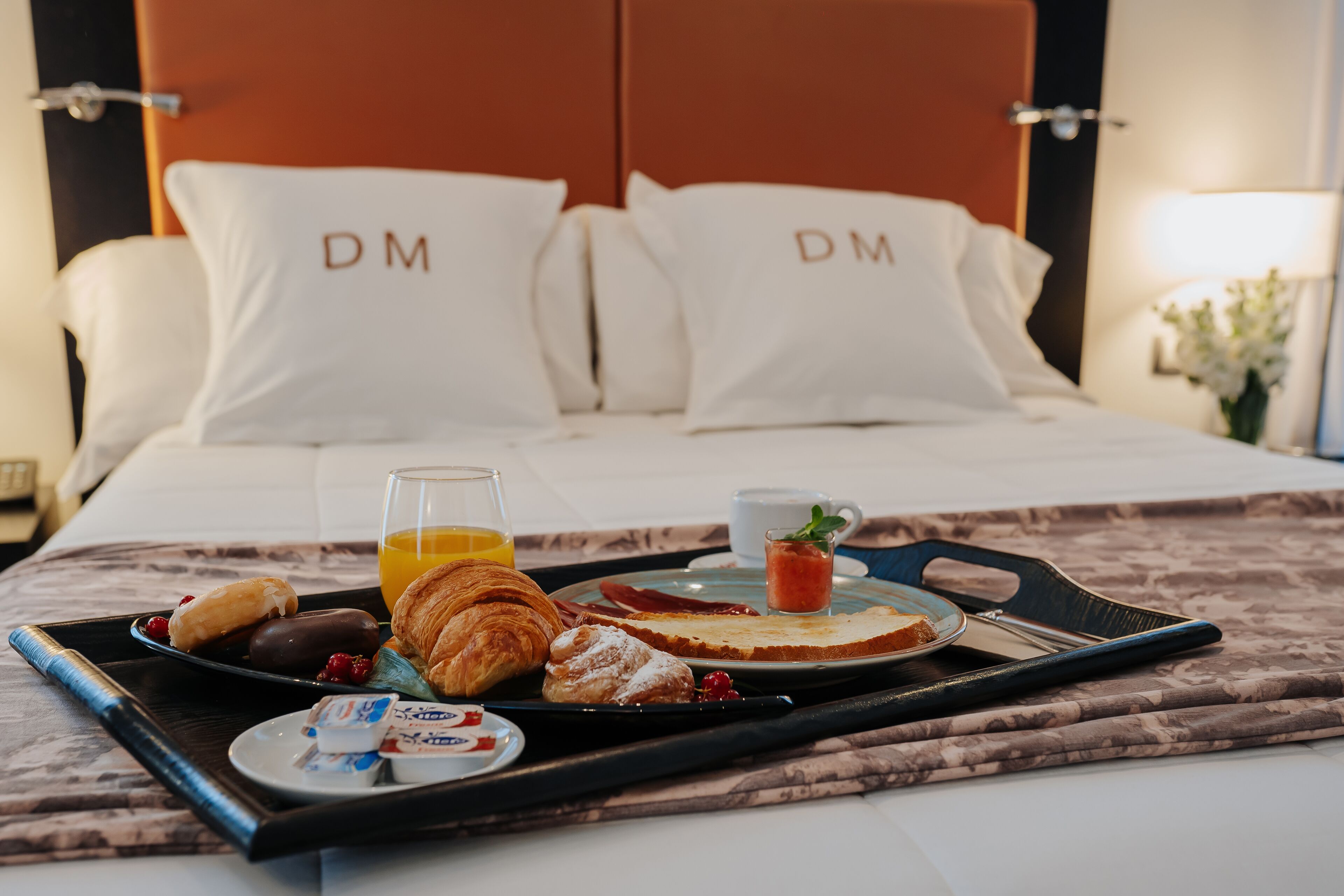 A neatly made breakfast tray with croissants, a glass of orange juice, and a cup of coffee on a bed with white pillows labeled "DM".