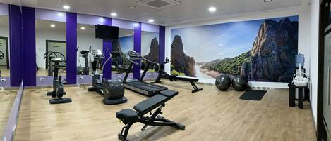 Un gimnasio con equipo de ejercicio, una banca, dispensador de agua y un mural de un paisaje montañoso.