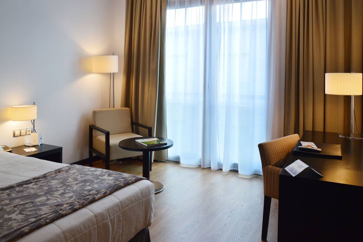A hotel room with a bed, two chairs, a small table, and a desk with a lamp.