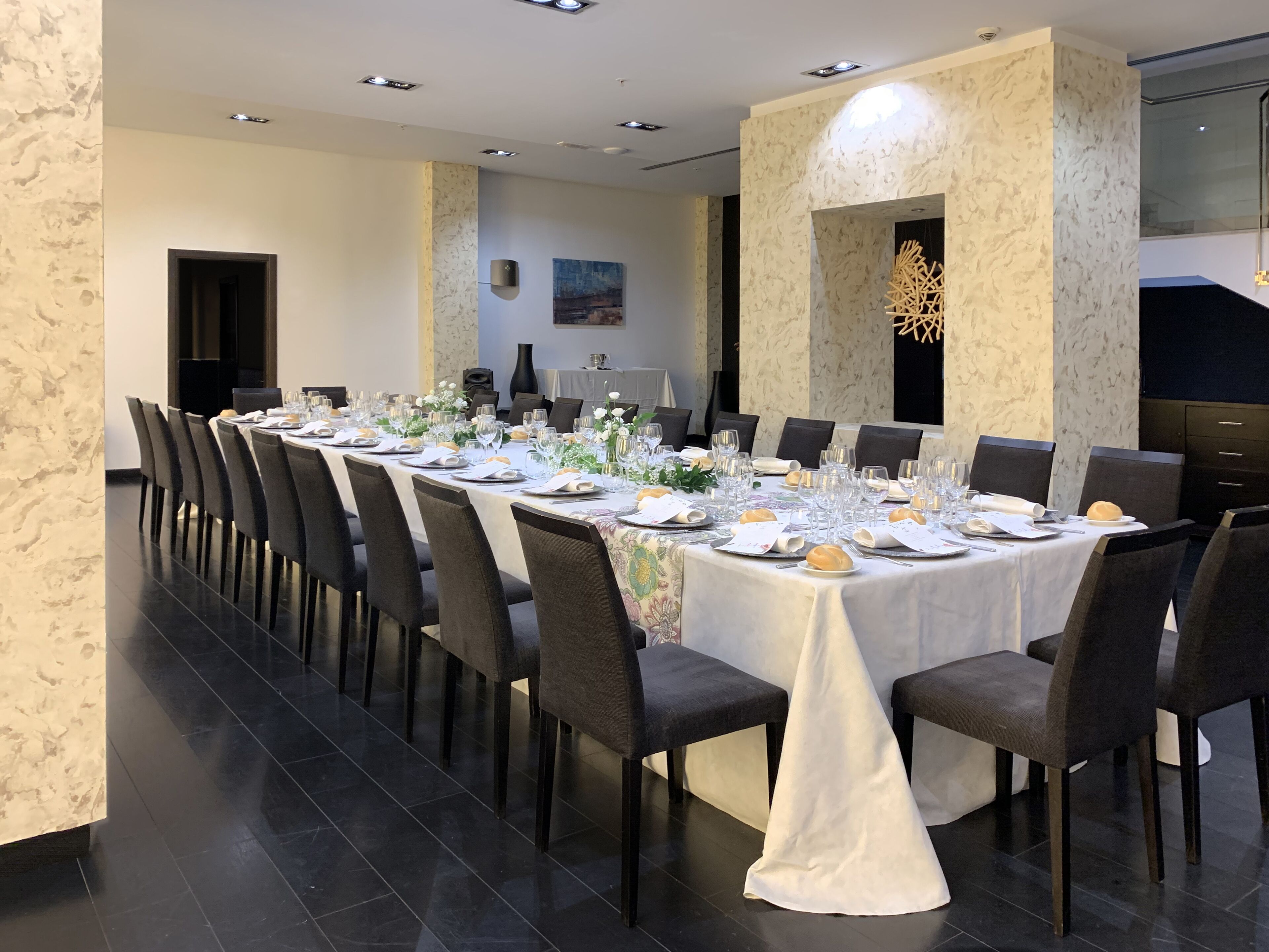 Una larga mesa de comedor preparada para una comida formal, con sillas, un florero con flores y un cuadro en la pared.