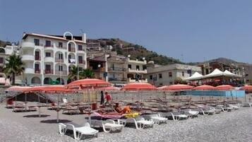 Una playa con sillas de descanso blancas y sombrillas rojas, un edificio con balcones y una colina al fondo.