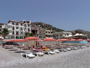 A beach with white lounge chairs and red umbrellas, a building with balconies, and a hillside in the background. - Albatros Beach Hotel - Letojanni (Letojanni)