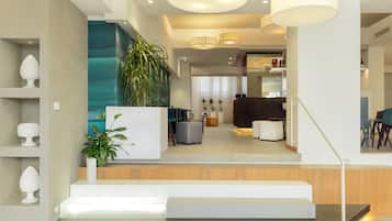 Un interior moderno con una escalera que conduce a una sala de estar, destacando un mostrador de recepción de madera, una planta grande y una zona de estar con un sofá oscuro y una mesa de centro.