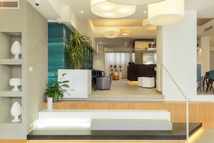 Un interior moderno con una escalera que conduce a una sala de estar, destacando un mostrador de recepción de madera, una planta grande y una zona de estar con un sofá oscuro y una mesa de centro.