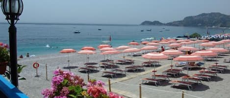A beach with rows of pink umbrellas and lounge chairs, a clear view of the sea, and a lamp post in the foreground.
