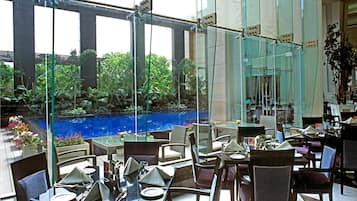 An indoor dining area with tables and chairs, a glass wall overlooking a garden and a pool.